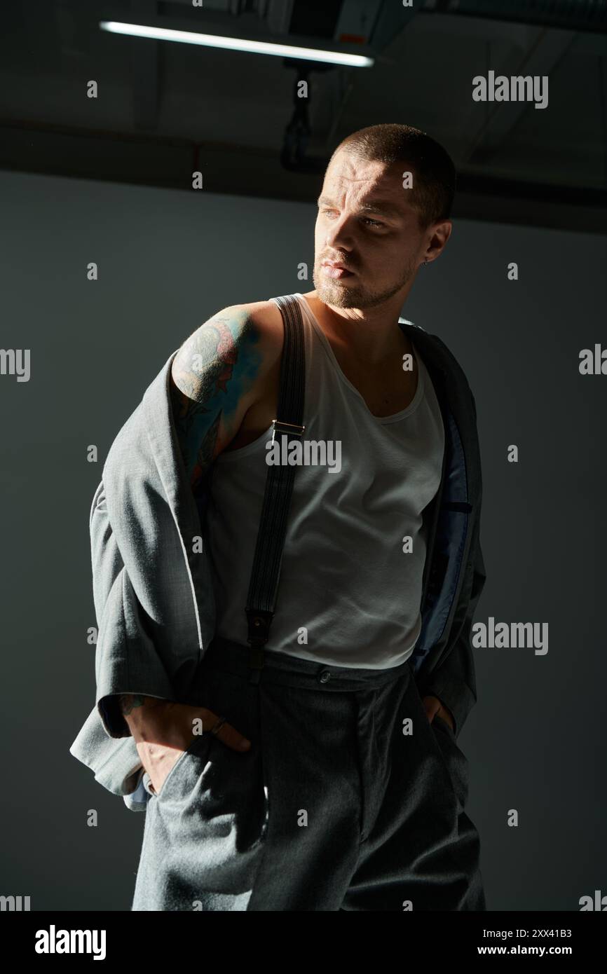 Uomo con taglio vivace e tatuaggi in studio, con canotta bianca, bretelle, giacca grigia. Foto Stock