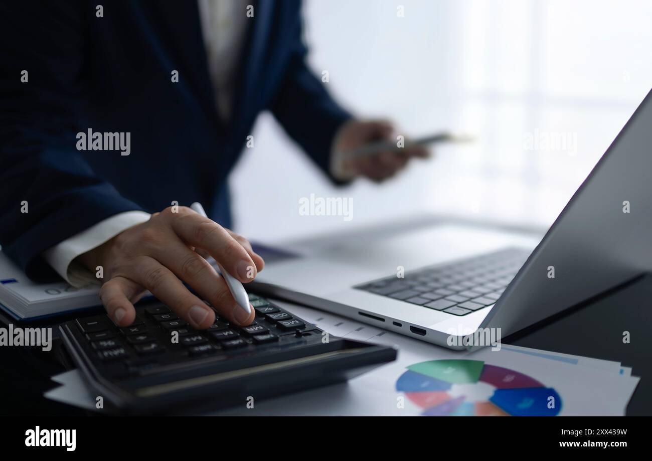 Un uomo d'affari è impegnato nell'analisi finanziaria e nella pianificazione aziendale utilizzando una calcolatrice e un laptop. L'immagine ritrae un ambiente professionale in cui Foto Stock