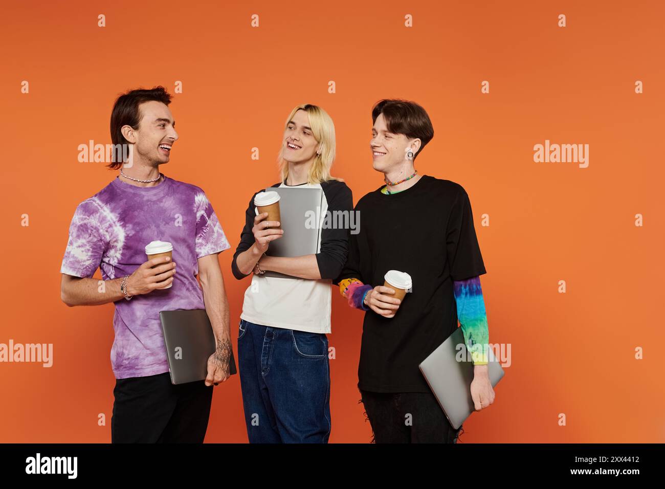 Tre giovani amici queer vestiti con un abbigliamento colorato si uniscono in uno studio con sfondo arancione. Foto Stock