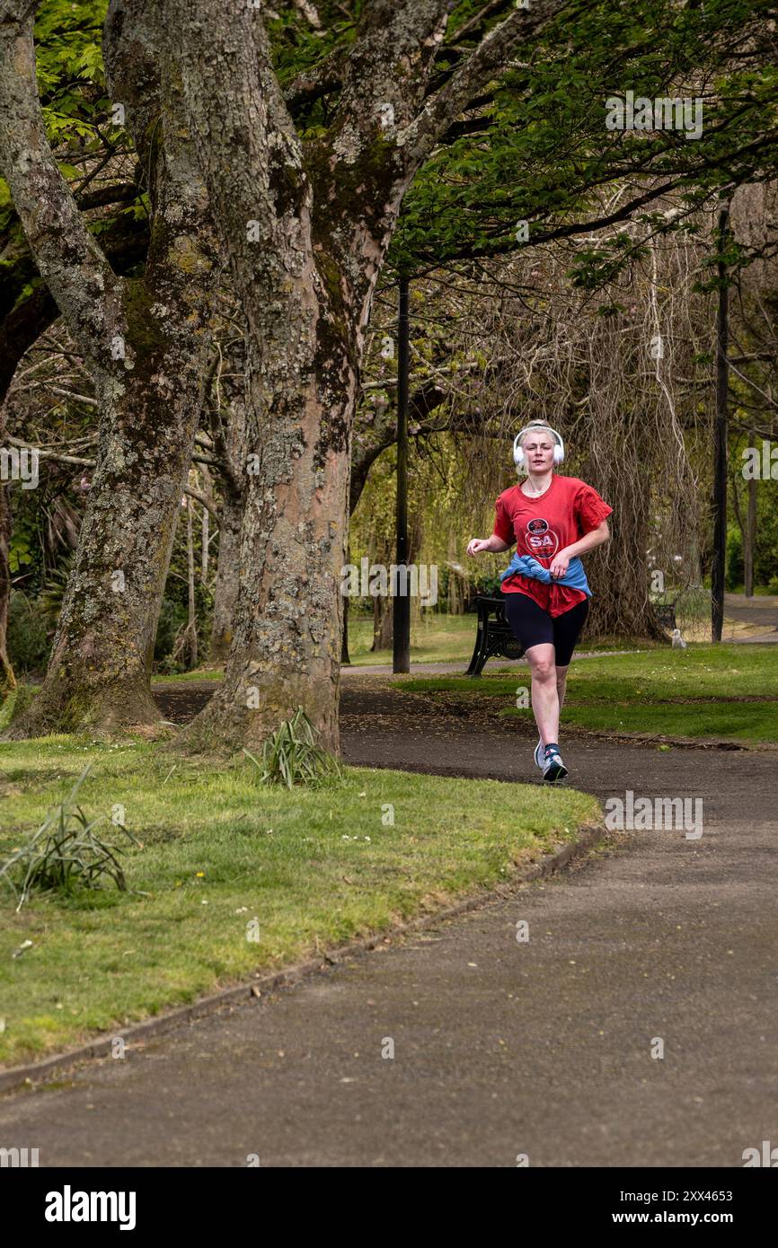 Una jogger runner che ascolta le sue cuffie mentre fa jogging attraverso i giardini Trenance di Newquay in Cornovaglia nel Regno Unito. Foto Stock