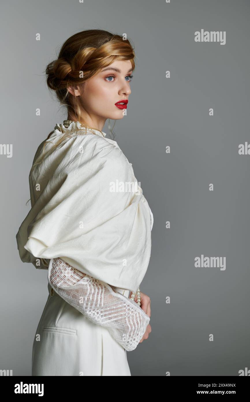 Una giovane donna con un elegante abbigliamento bianco si staglia su uno sfondo grigio, il suo sguardo è rivolto lateralmente. Foto Stock