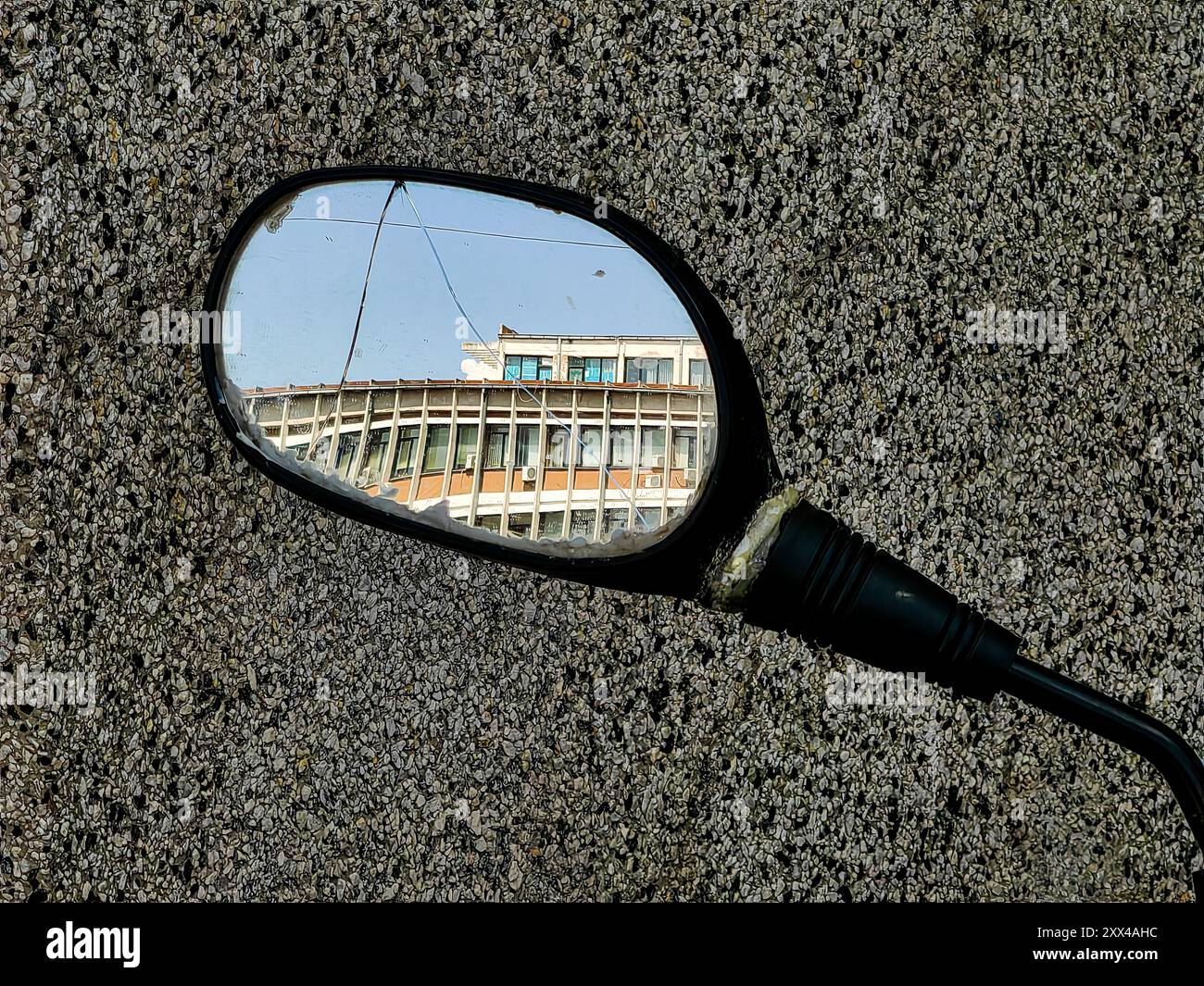 Riflessioni del passato: Nello specchio danneggiato di vista laterale di uno scooter che riflette un vecchio edificio ospedaliero contro un muro di granito - Sunny Day with Blue Sky Foto Stock