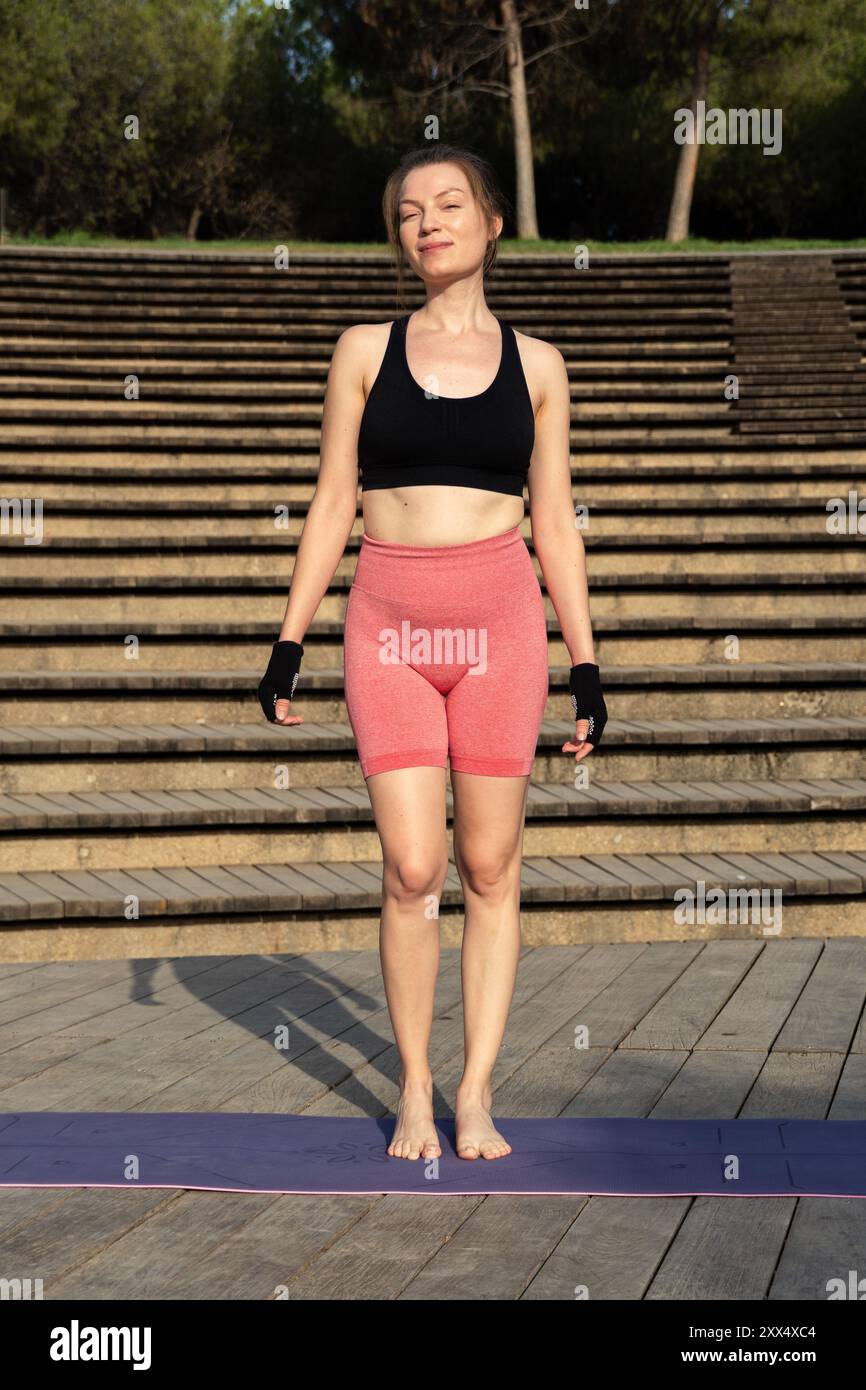 Una giovane donna rossa, vestita con abbigliamento atletico e guanti, si erge su un ponte di legno illuminato dal sole, sorridente e pronta per la sua lezione di yoga all'aperto di Pilates, Foto Stock