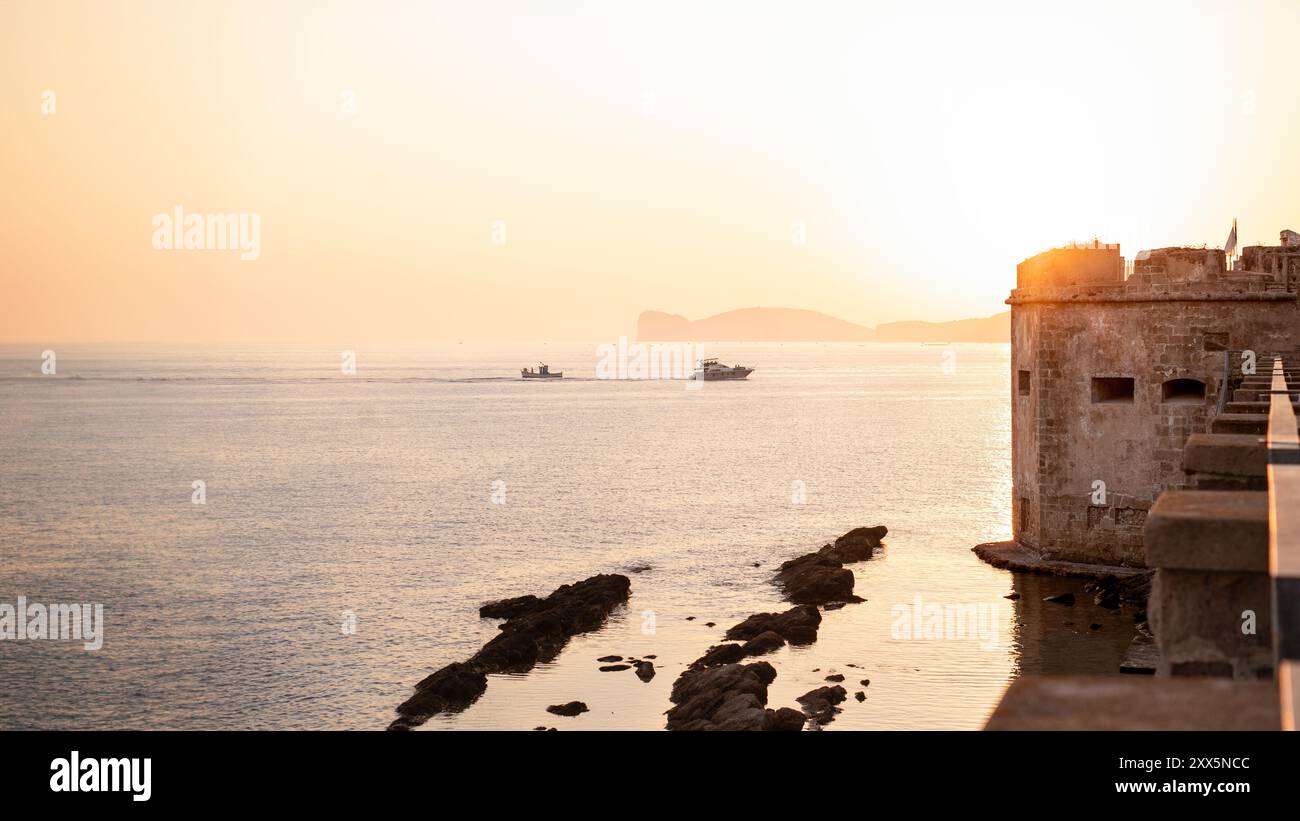 Vista parziale dell'antica cinta muraria difensiva della città costiera di Alghero di fronte ad un intenso tramonto sul mare con due barche che ritornano al p Foto Stock