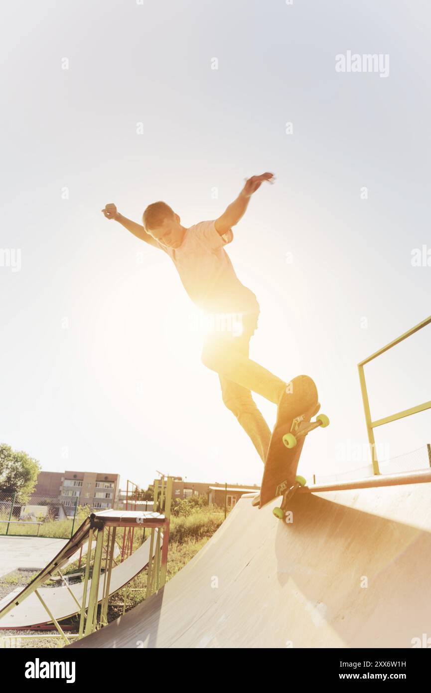 Il pattinatore teenager è appeso su una rampa su uno skateboard in uno skate Park al tramonto. Grandangolo. Immagine calda e soleggiata Foto Stock