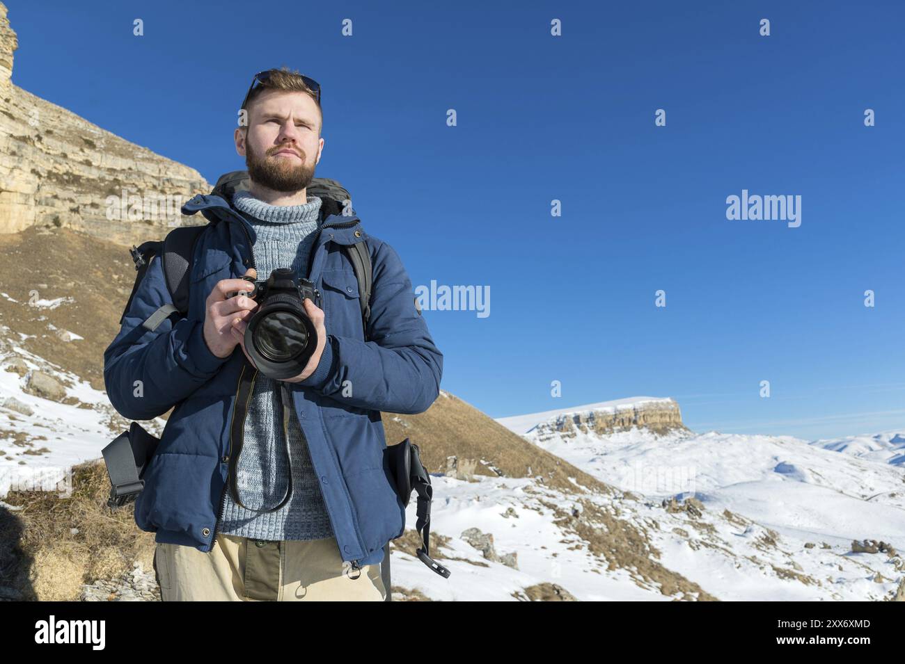 Ritratti Un fotografo hipster barbuto con uno zaino e indossa occhiali da sole con piedistalli con una fotocamera DSLR in mano sullo sfondo di e. Foto Stock