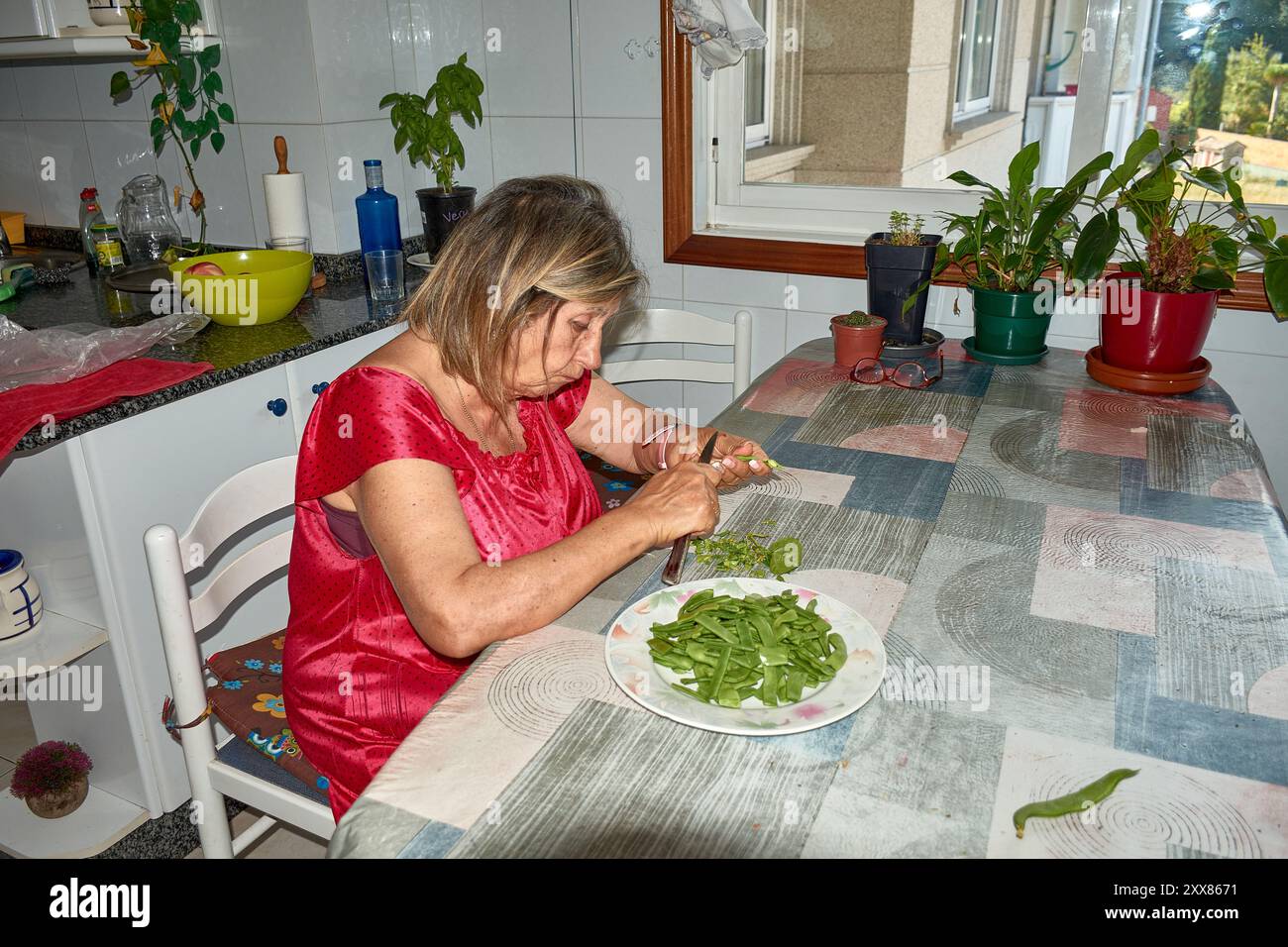 donna anziana seduta al suo tavolo della cucina, affettando con cura i fagiolini con un coltello. La scena riflette la semplicità e il comfort della vita quotidiana, wh Foto Stock