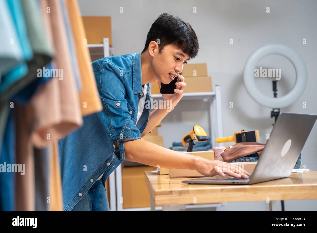 Giovane venditore online felice che parla al telefono con il cliente mentre controlla gli ordini in arrivo dal suo computer portatile Foto Stock