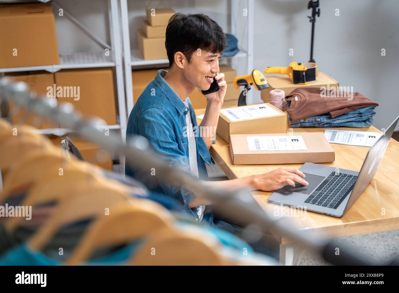 Giovane venditore online felice che parla al telefono con il cliente mentre controlla gli ordini in arrivo dal suo computer portatile Foto Stock