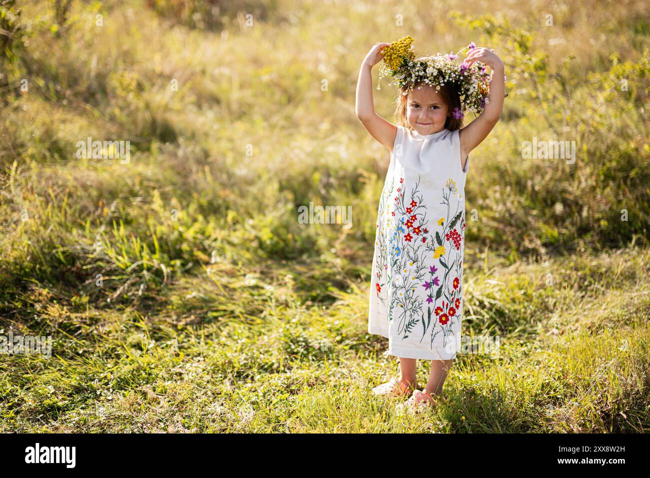 Adorabile ragazza che indossa un abito floreale e una corona di fiori, in piedi in un prato illuminato dal sole, irradiando gioia e innocenza. Perfetto per i concetti di bambino Foto Stock