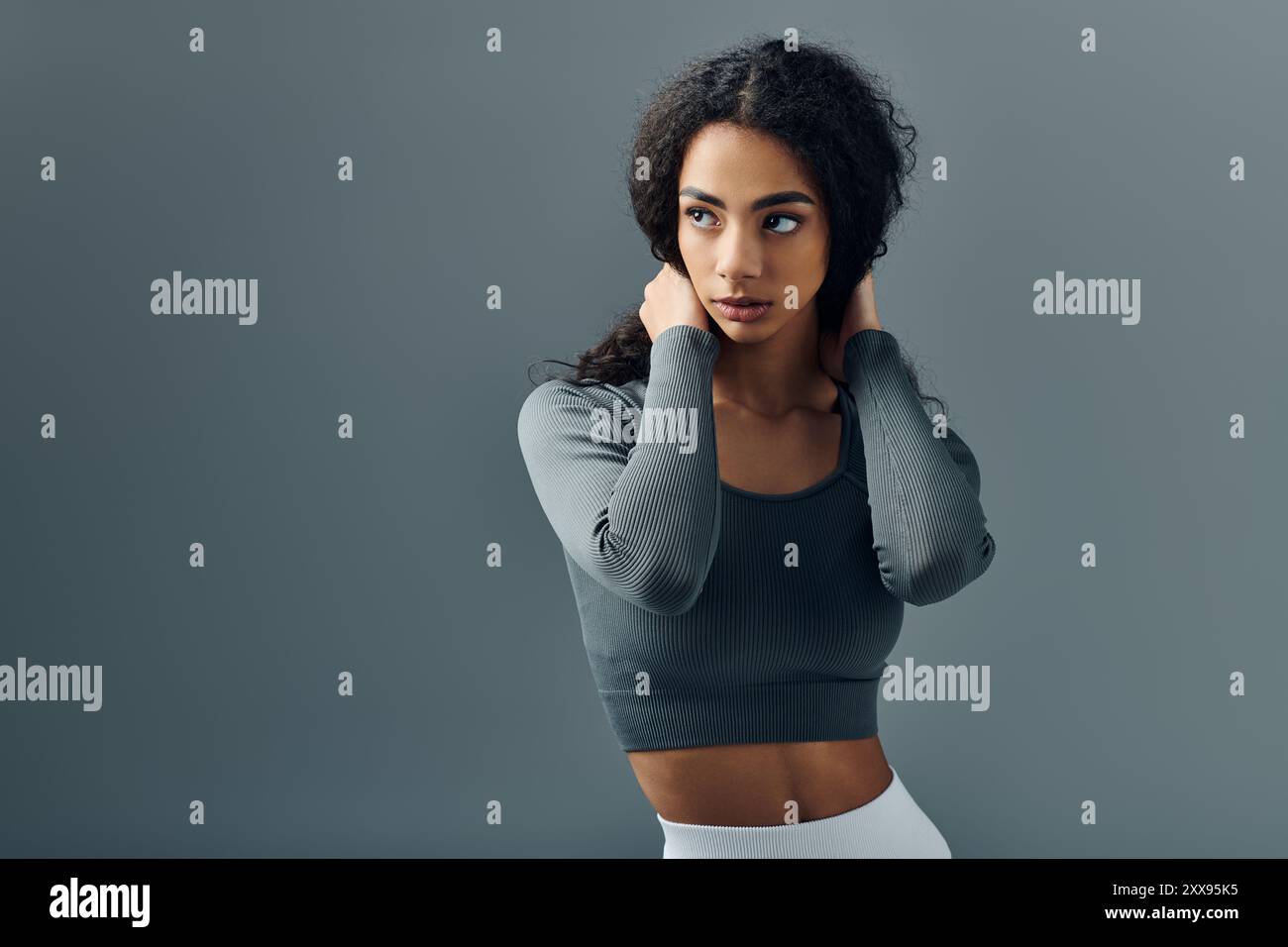Una giovane donna con la parte superiore grigia si erge davanti a uno sfondo grigio, guardando lateralmente e regolando i capelli. Foto Stock