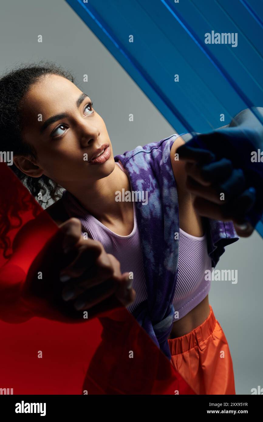 Una giovane donna afroamericana in abbigliamento sportivo si estende verso pannelli di vetro colorati in uno studio, con gli occhi che guardano verso l'alto. Foto Stock