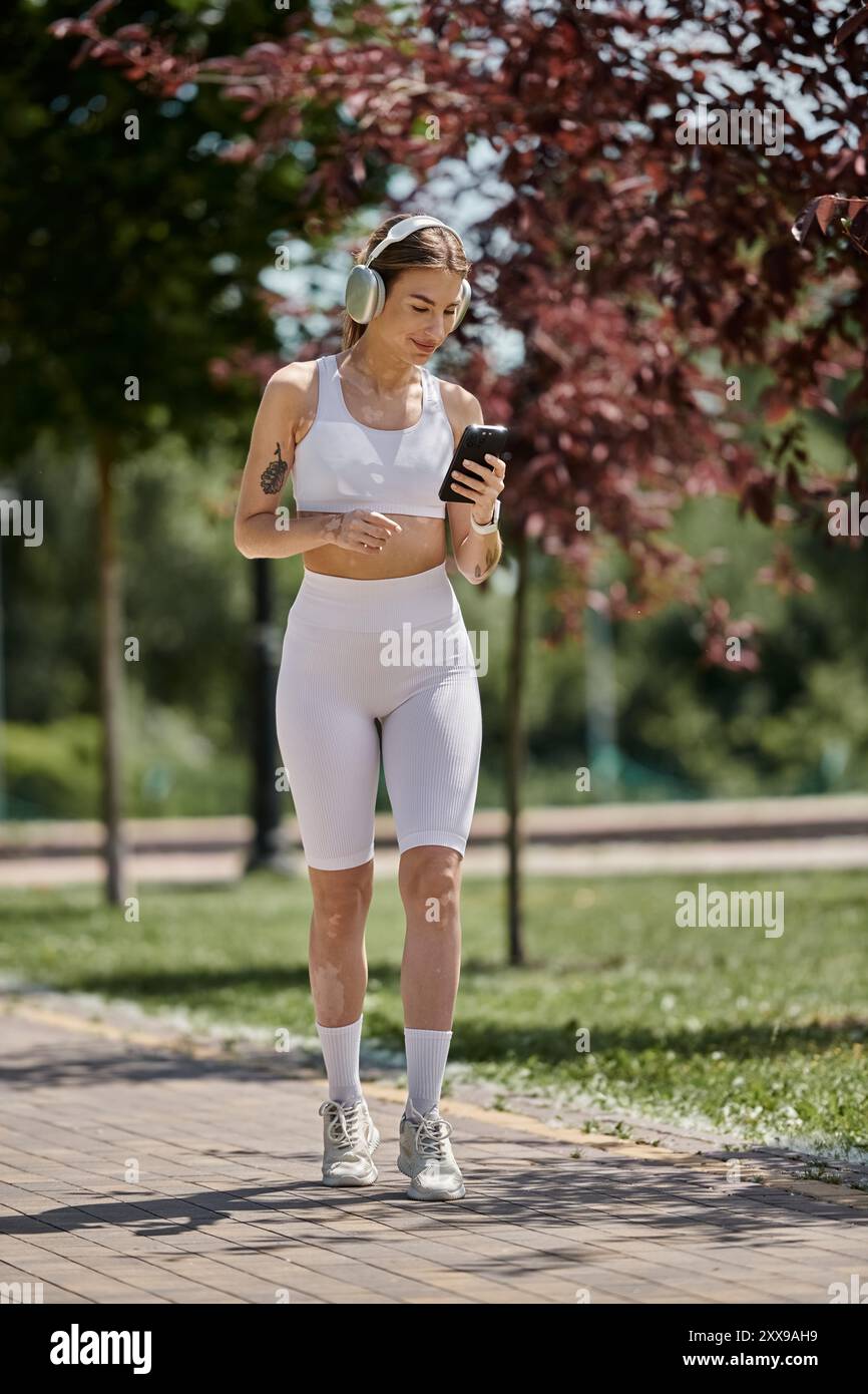 Una giovane donna con vitiligine cammina in un parco, indossando abiti da ginnastica bianchi e cuffie, mentre guarda il suo telefono. Foto Stock