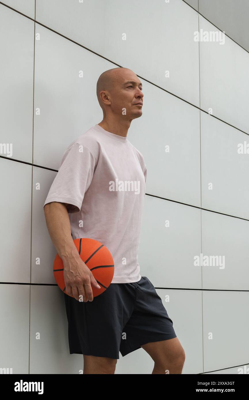 Ritratto di un bellissimo uomo adulto sportivo con t-shirt bianca che regge la palla arancione da basket e guarda lontano vicino al muro bianco. Foto Stock