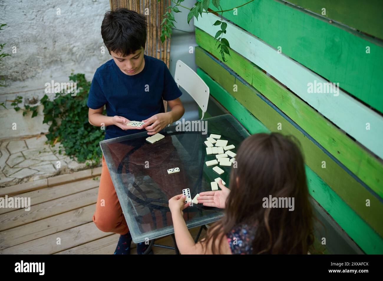 Due bambini che giocano a domino all'esterno in un giardino, circondato da pannelli verdi e piante. Si concentrano sul gioco, godendosi un pomeriggio rilassato. Foto Stock