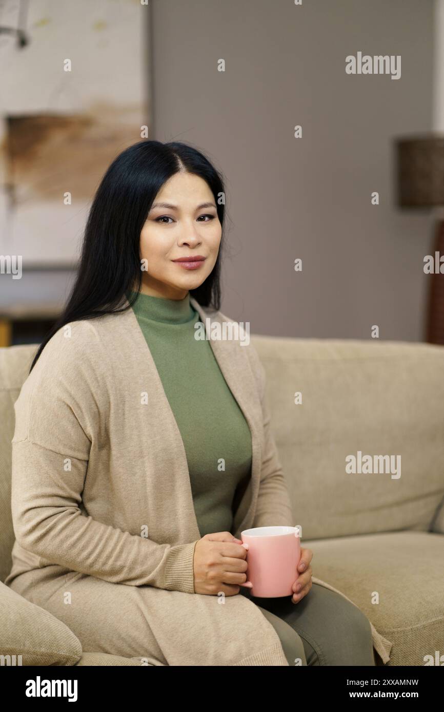 Una donna è seduta su un divano tenendo in mano una tazza rosa. Indossa un maglione verde e una giacca abbronzante Foto Stock