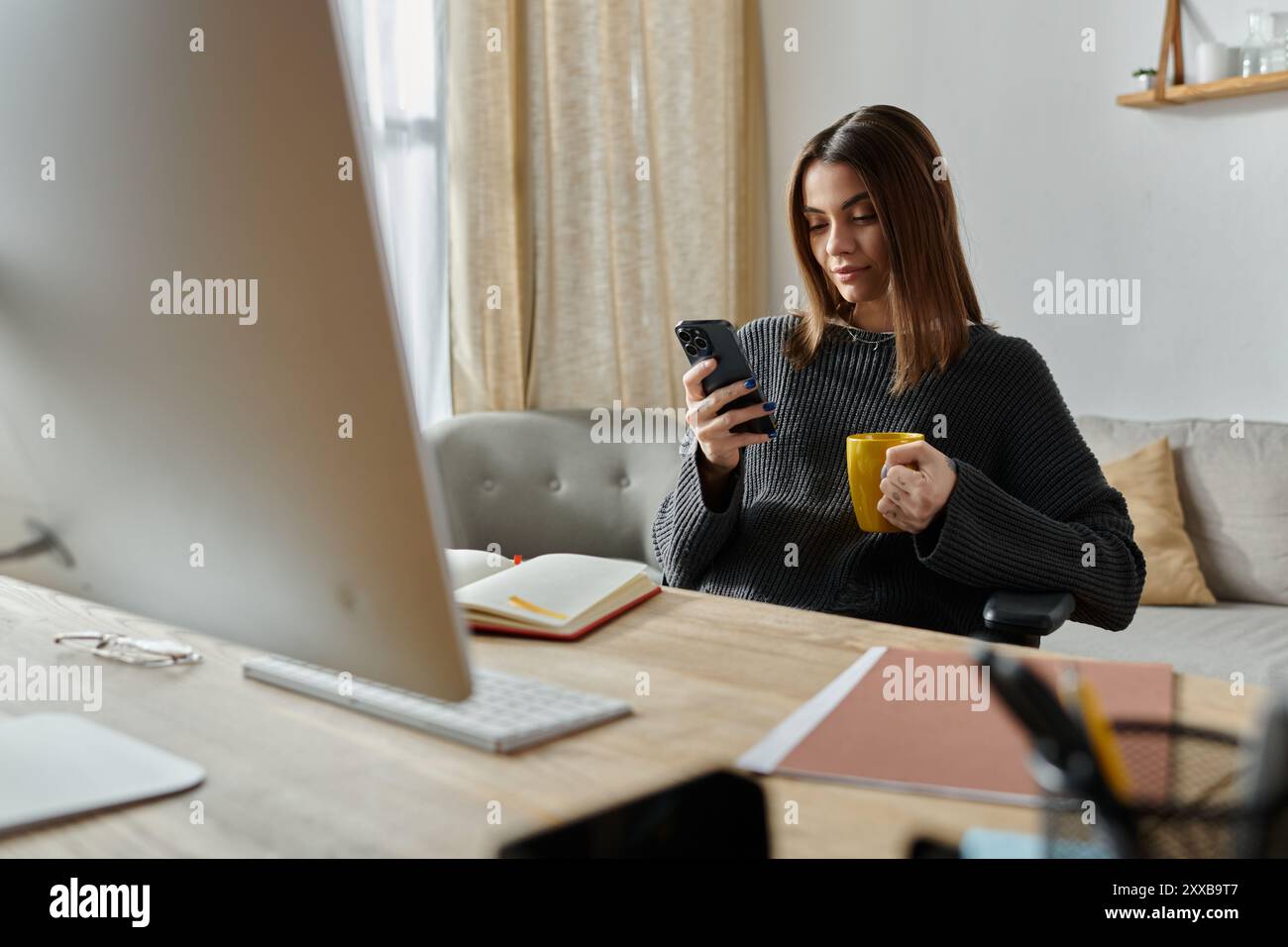 Una donna lavora da casa, incollata al telefono, sorseggiando un caffè, probabilmente gestendo il marketing digitale. Foto Stock