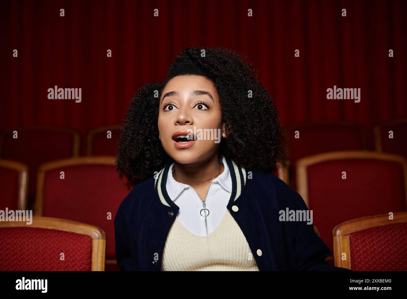 Una giovane donna con una giacca blu scuro siede in un cinema, guardando lo schermo con un'espressione sconvolgente. Foto Stock