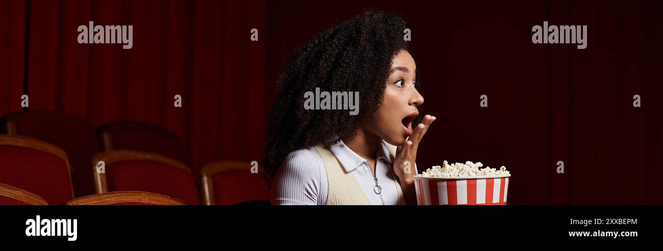 Una giovane donna afroamericana con i capelli ricci siede in un cinema, reagisce con sorpresa e tiene in mano un secchio di popcorn. Foto Stock