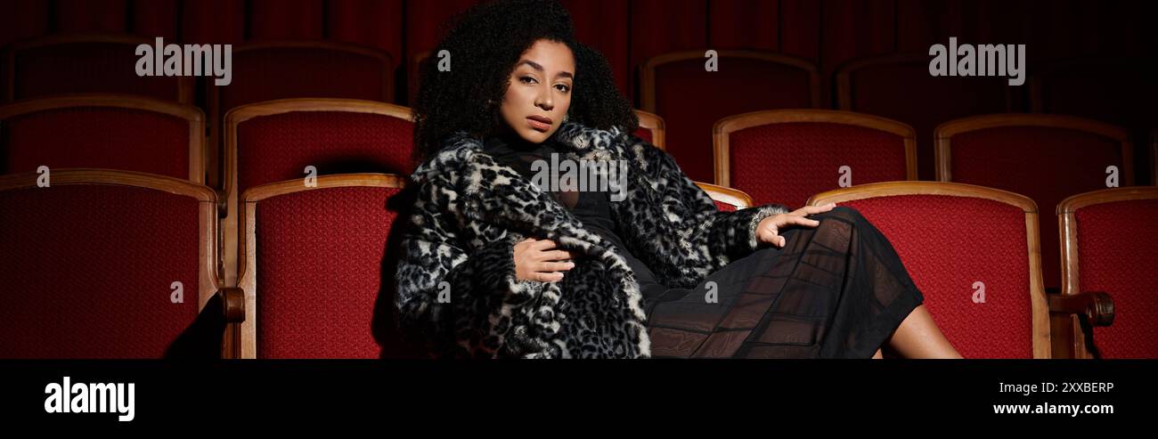 Un'elegante donna afro-americana con un cappotto alla moda siede in un posto a teatro, guardando un film con un'espressione mirata. Foto Stock