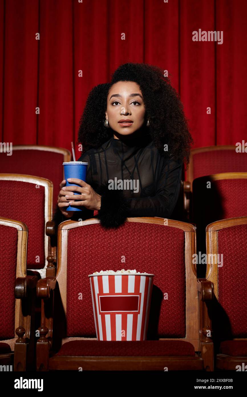 Una donna elegante siede in un cinema, sorseggiando un drink e guardando lo schermo, la sua espressione piena di intrighi. Foto Stock