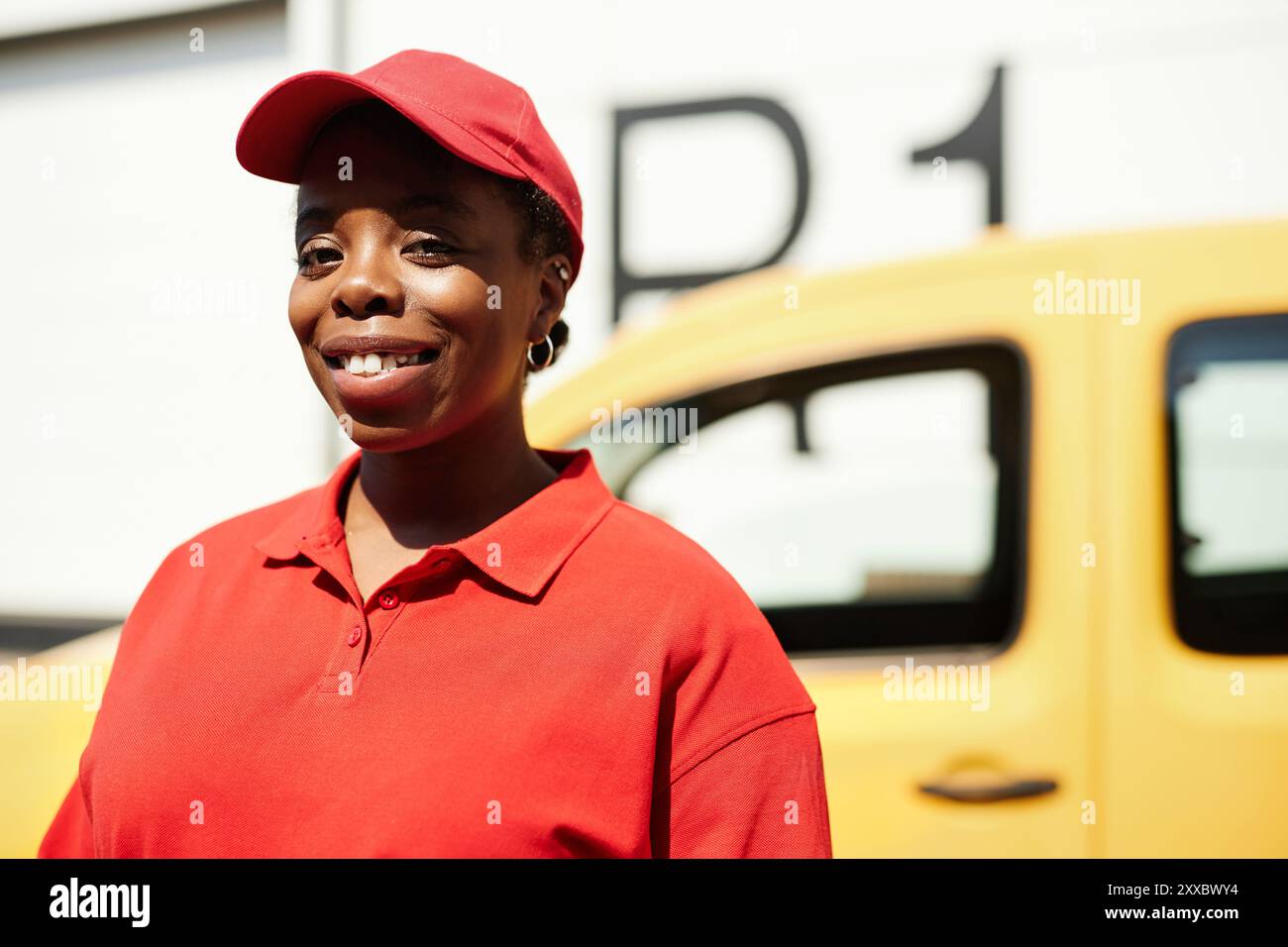 Donna afroamericana sorridente mentre si trova accanto al furgone giallo nel parcheggio. Indossa un berretto rosso e una polo, trasudando sicurezza e felicità Foto Stock