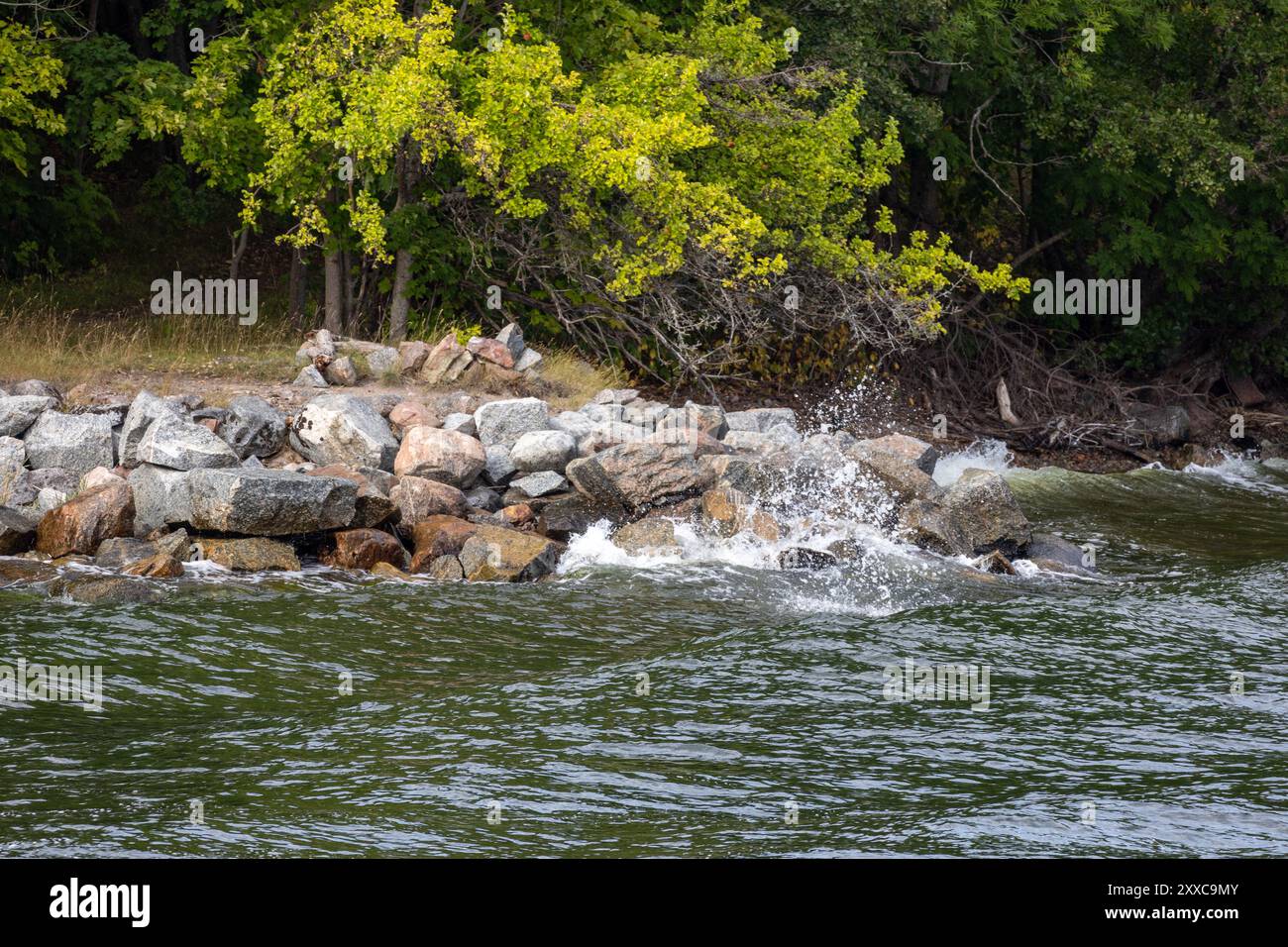 Un litorale roccioso con onde che spruzzano contro le pietre, circondato da lussureggianti alberi verdi. L'acqua è calma con morbide increspature, creando un ambiente sereno Foto Stock