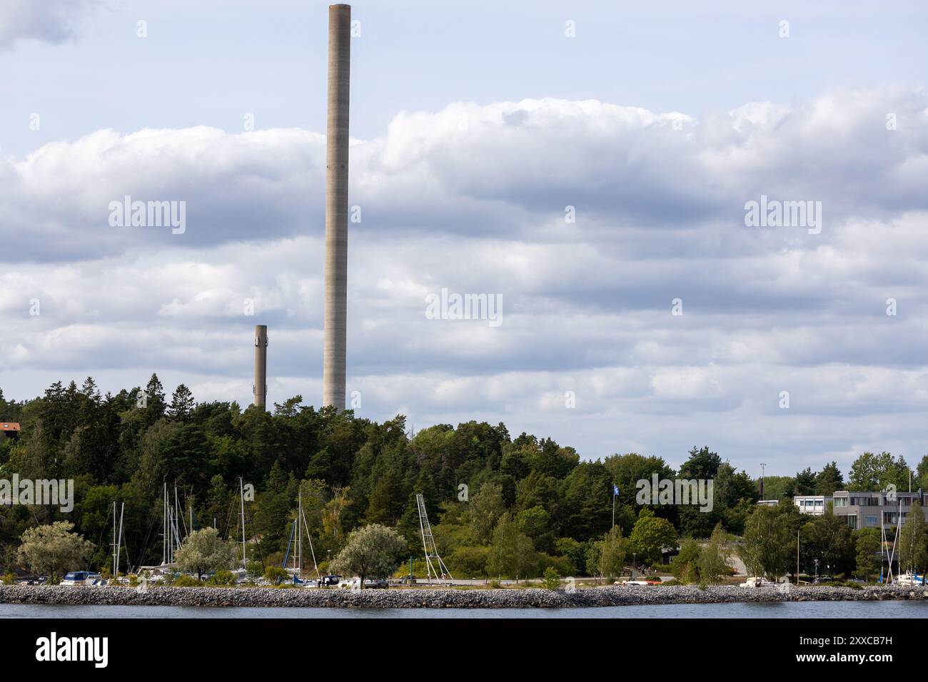 Una vista panoramica di un fronte mare con alti fumogeni sullo sfondo. Il primo piano presenta un porticciolo con diverse barche a vela e lussureggianti alberi verdi. T Foto Stock