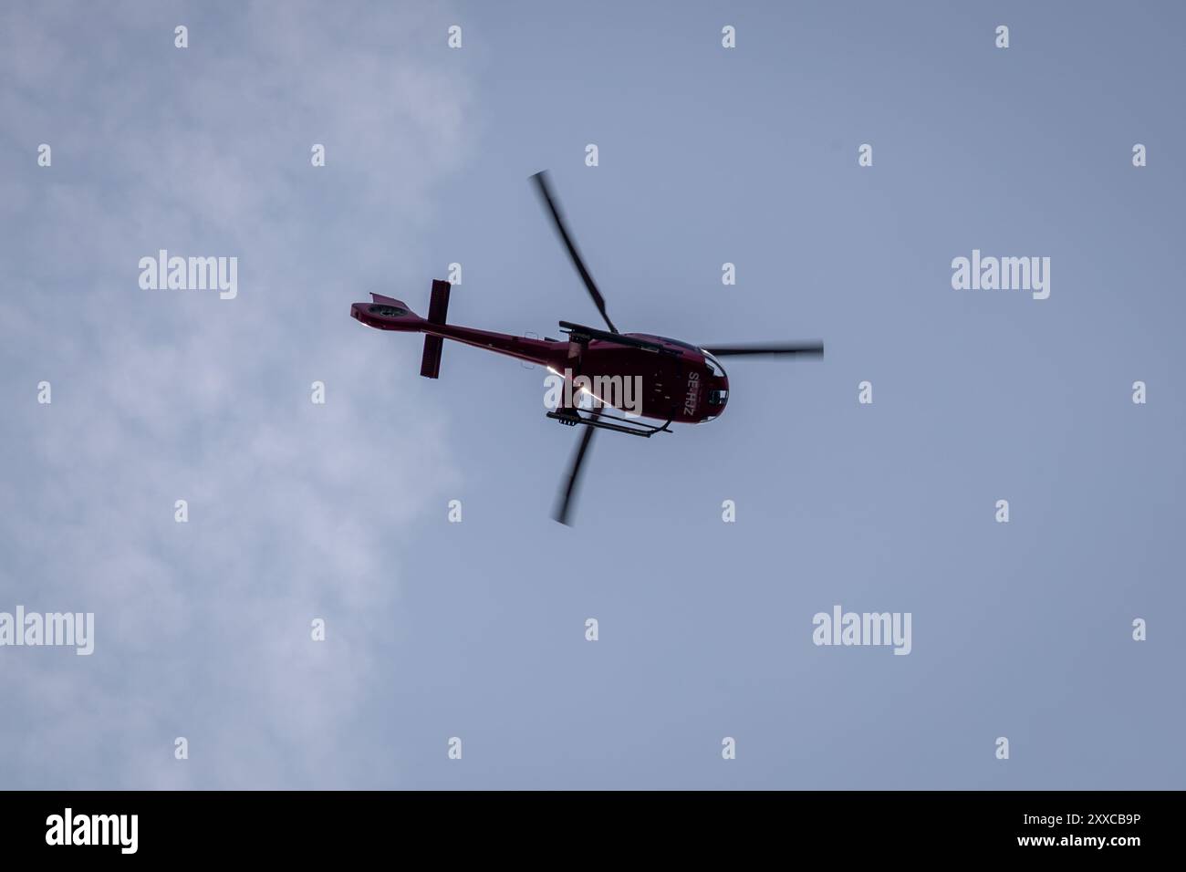 Un elicottero rosso che vola contro un cielo blu con nuvole sparse. L'elicottero è visto dal basso, mostrando le pale del rotore in movimento. Foto Stock