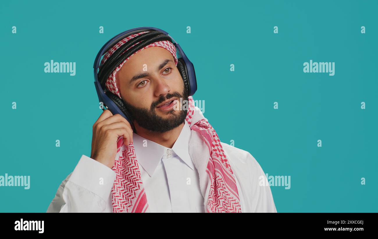 Un uomo che balla con la fotocamera, ascolta musica moderna con le cuffie e divertiti in studio. Uomo mediorientale in abbigliamento tradizionale che mostra movimenti di danza funky e canti. Foto Stock