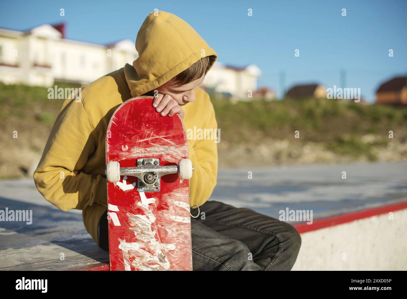 Primo piano di un giovane hipster vestito con una felpa e jeans seduti tristi in uno skate Park e con in mano uno skateboard Foto Stock