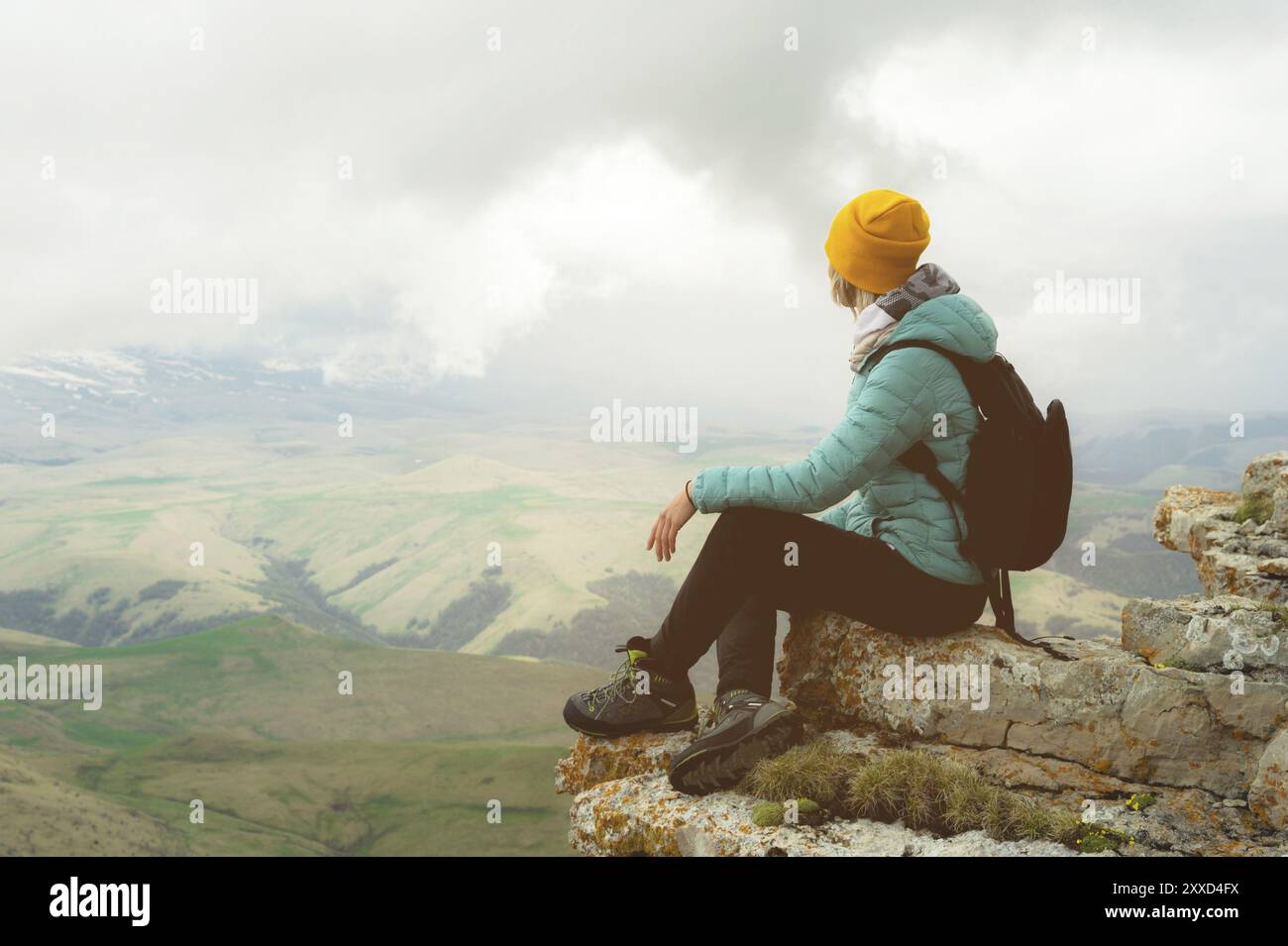 Giovane donna con uno zaino che si siede sul bordo di una roccia e guarda il cielo con le nuvole Foto Stock