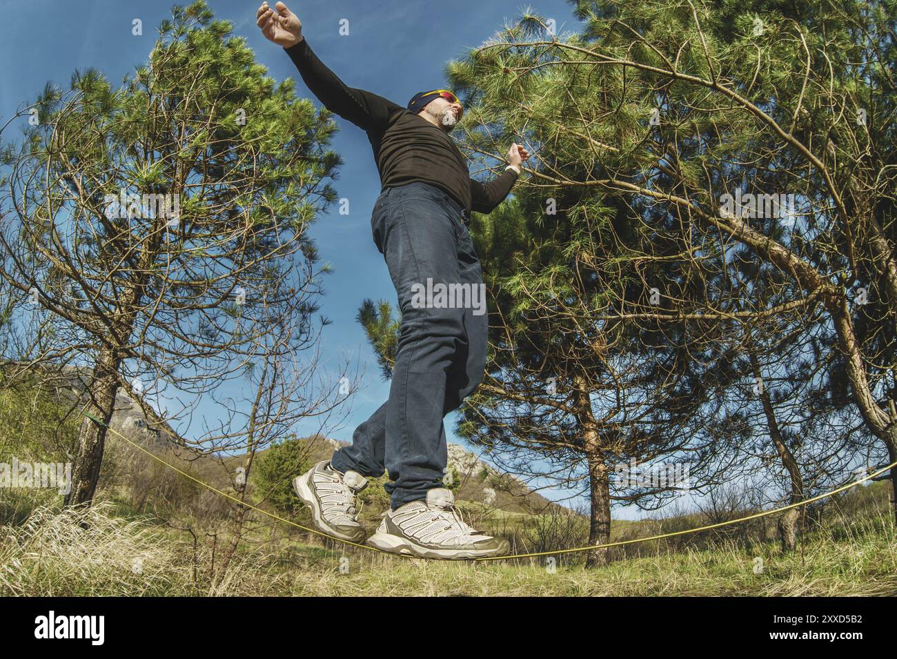 Un uomo, invecchiato con la barba e con gli occhiali da sole, si bilancia su una linea lenta all'aria aperta tra due alberi al tramonto sul cielo blu di sfondo Foto Stock