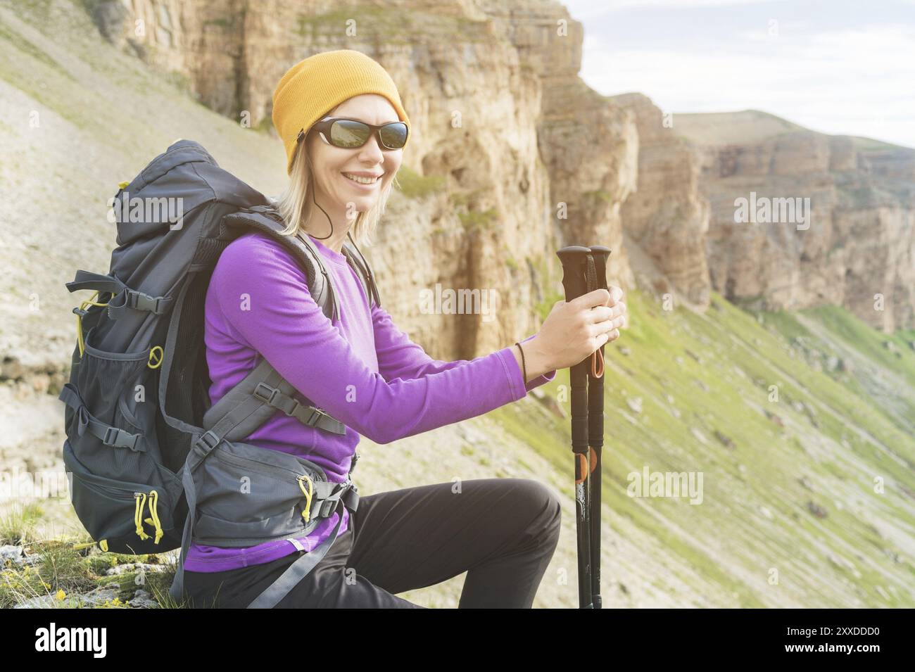 Viaggiatrice sorridente con un cappello giallo e un paio di occhiali da sole che si bagliano ai piedi di rocce epiche con uno zaino e guardano in macchina fotografica Foto Stock
