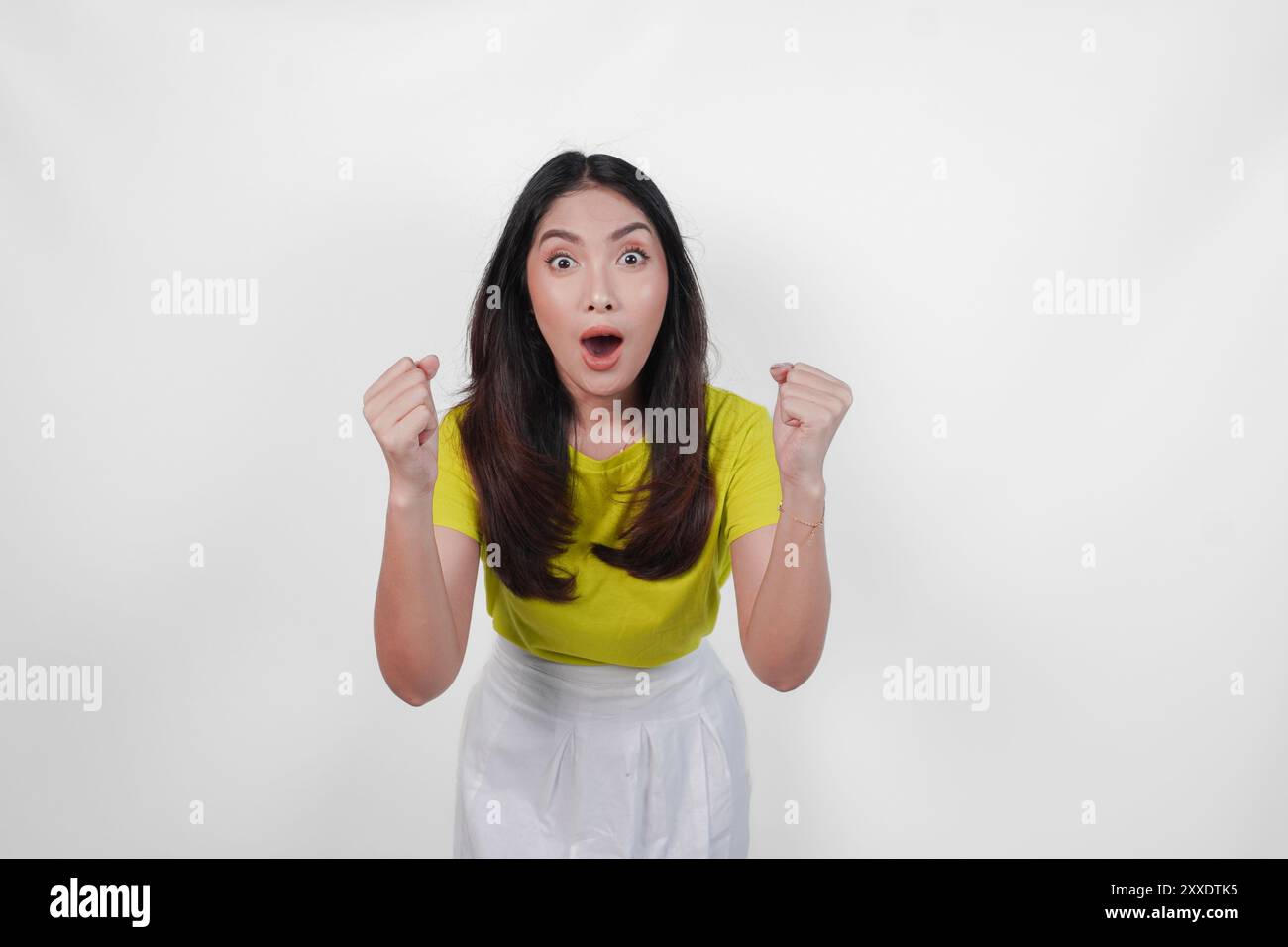 Allegra donna asiatica con una camicia gialla e una gonna bianca sorride e solleva le braccia in aria. Concetto di gioia ed eccitazione. Foto Stock