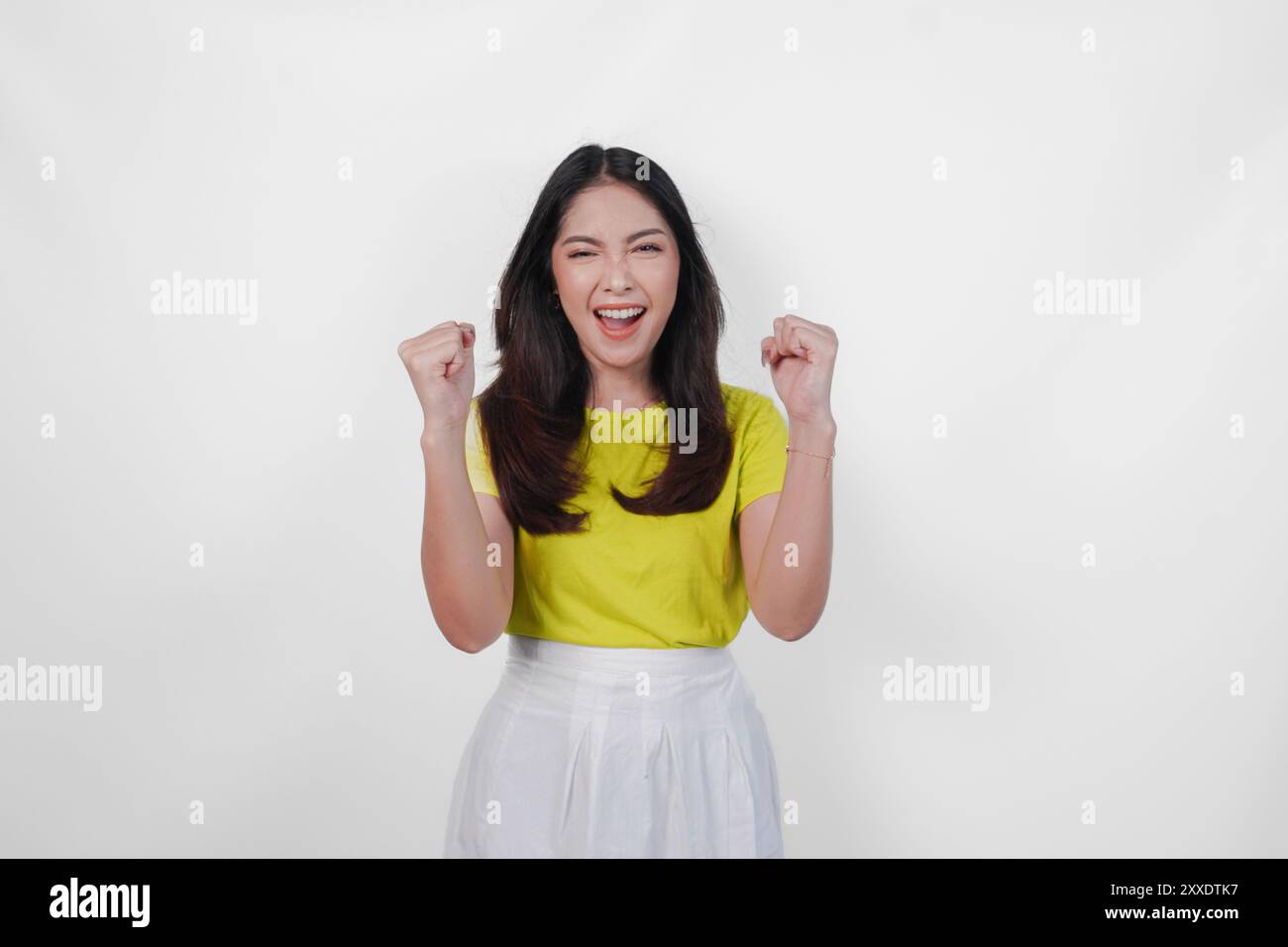 Allegra donna asiatica con una camicia gialla e una gonna bianca sorride e solleva le braccia in aria. Concetto di gioia ed eccitazione. Foto Stock