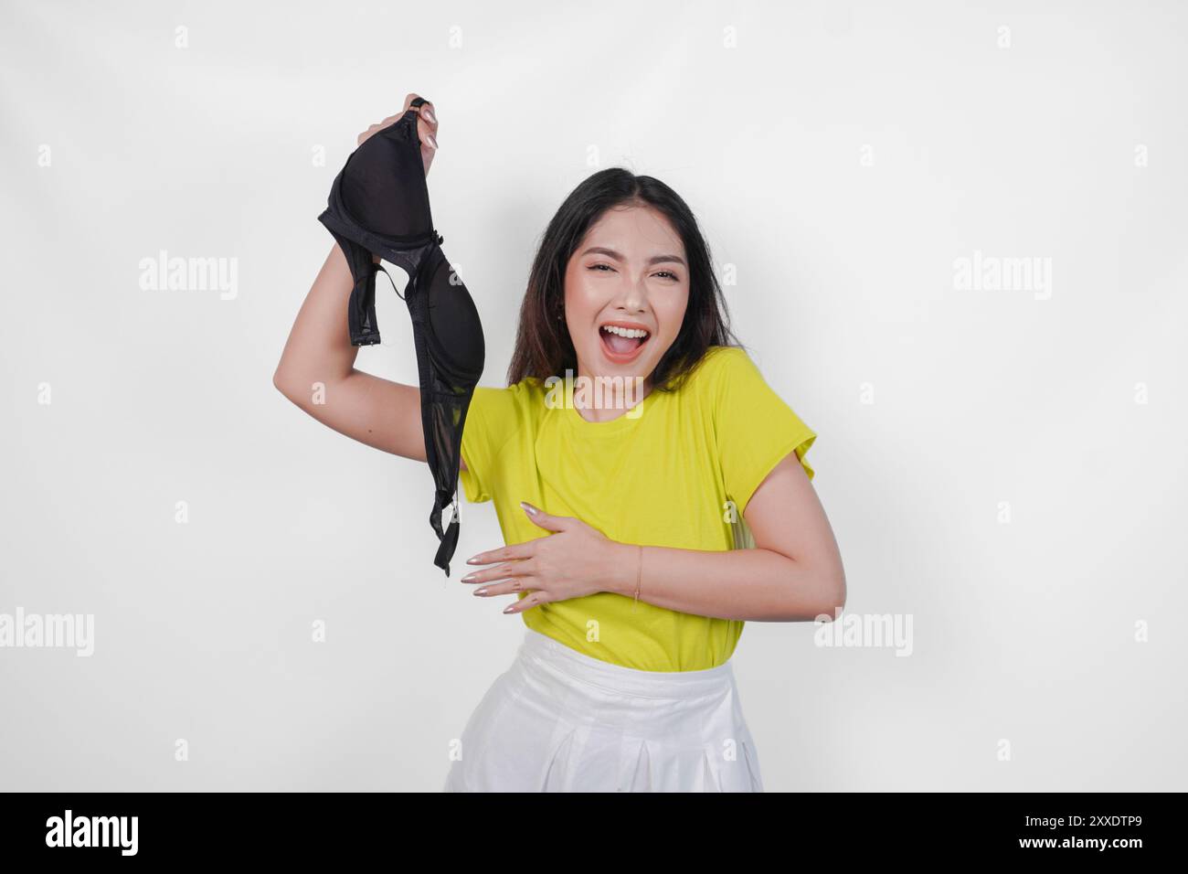 Sorpresa giovane donna asiatica in maglia gialla sta tenendo in mano un reggiseno nero per la festa del giorno del reggiseno, isolato da sfondo bianco. Foto Stock