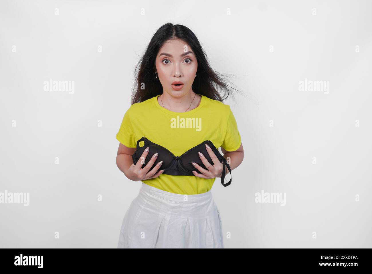La giovane donna asiatica in maglietta gialla tiene un reggiseno nero sul petto per festeggiare il giorno del reggiseno con un'espressione scioccata, isolata da sfondo bianco Foto Stock
