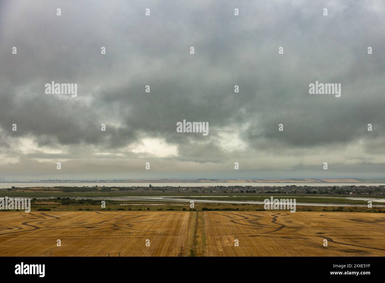 Castello di Hadleigh, Regno Unito. 24 agosto 2024. Una vista grandangolare di un campo agricolo raccolto di recente nell'Essex, Inghilterra, con binari visibili sulla superficie dorata-marrone. Oltre il campo si trova un estuario o un fiume, con una città lontana all'orizzonte. Il cielo è pieno di nuvole pesanti e scure, creando un drammatico contrasto tra terra e atmosfera. Un inizio bagnato e cupo per il fine settimana festivo al Castello di Hadleigh, affacciato sull'estuario del Tamigi. Penelope Barritt/Alamy Live News Foto Stock
