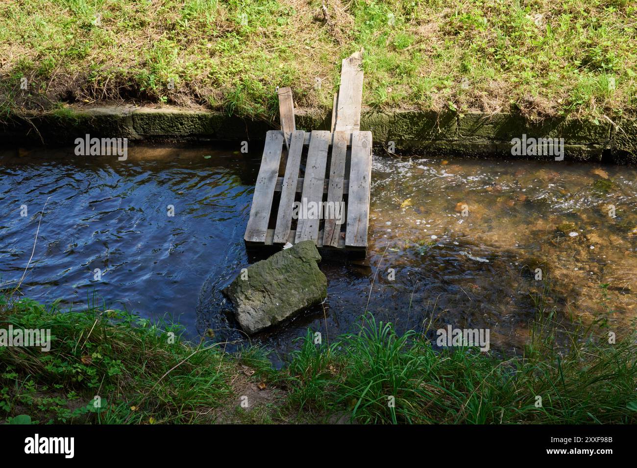 Improvvisatore, provisorischer Fussgängersteg über einen kleinen kanalisierten Bach aus einer Palette und einem großen Stein Foto Stock