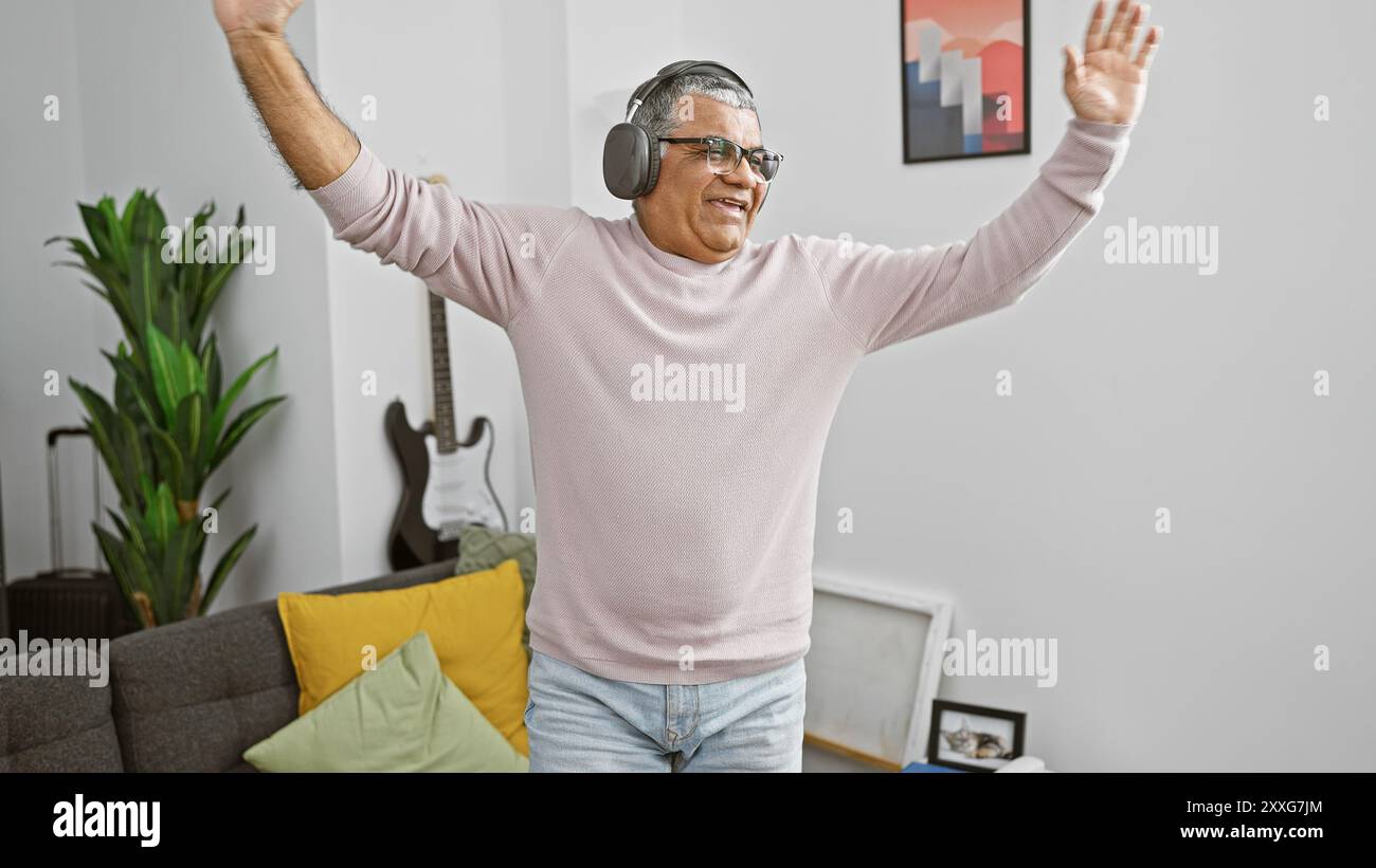 Un uomo allegro e maturo balla gioiosamente a casa con le cuffie, esprimendo felicità in un accogliente ambiente da salotto. Foto Stock