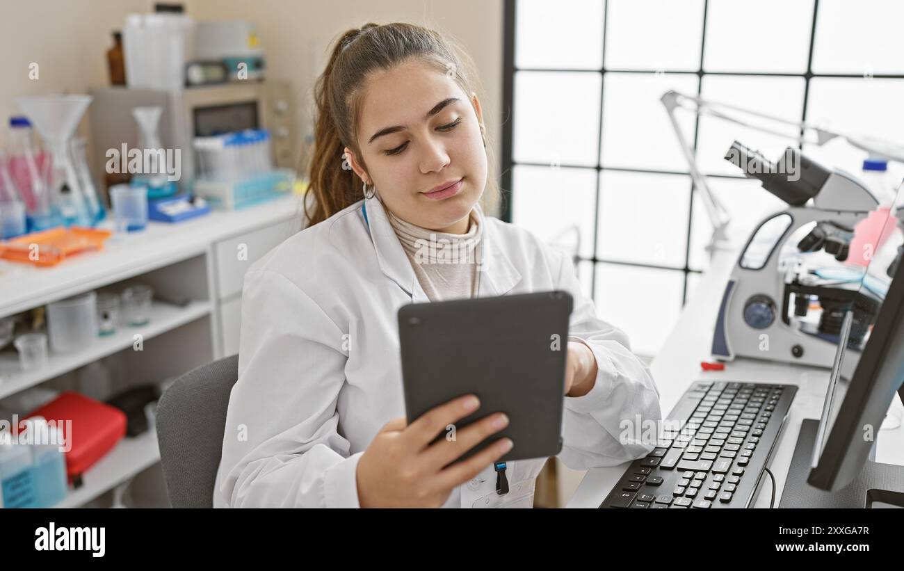 Una giovane scienziata ispanica analizza i dati su un tablet in un ambiente di laboratorio, rappresentando la tecnologia e l'assistenza sanitaria. Foto Stock