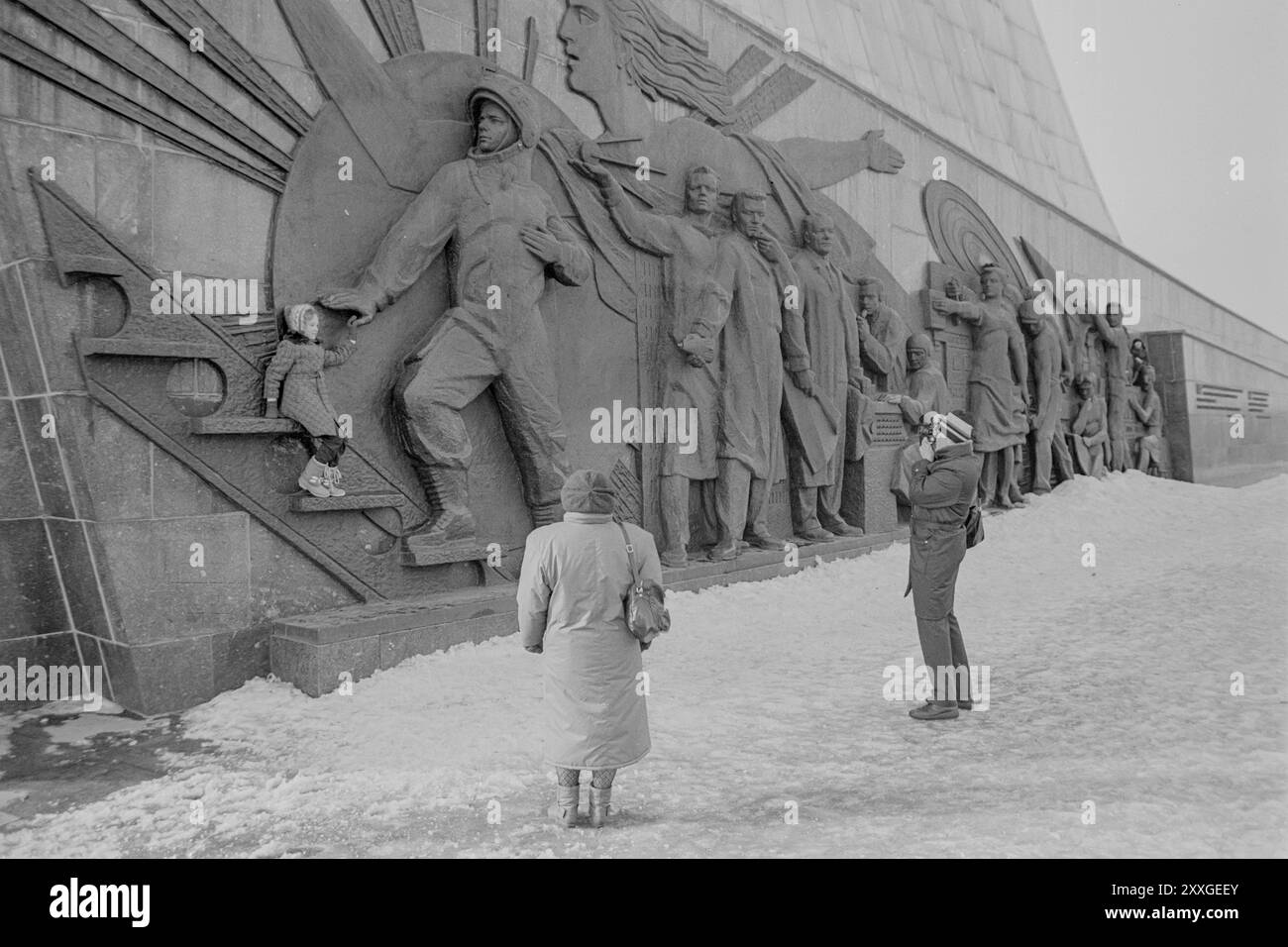 Un uomo scatta una foto di un bambino alla base del Monumento ai conquistatori dello spazio mentre una donna guarda. Il monumento, nella Mostra dei risultati dell'economia nazionale di Mosca (VDNKh), è stato costruito nel 1964 e supporta un obelisco alto 350 piedi sormontato da un veicolo spaziale. I rilievi raffigurano gli uomini e le donne che hanno lavorato al programma spaziale sovietico, tra cui un cosmonauta, scienziati e ingegneri. Foto Stock