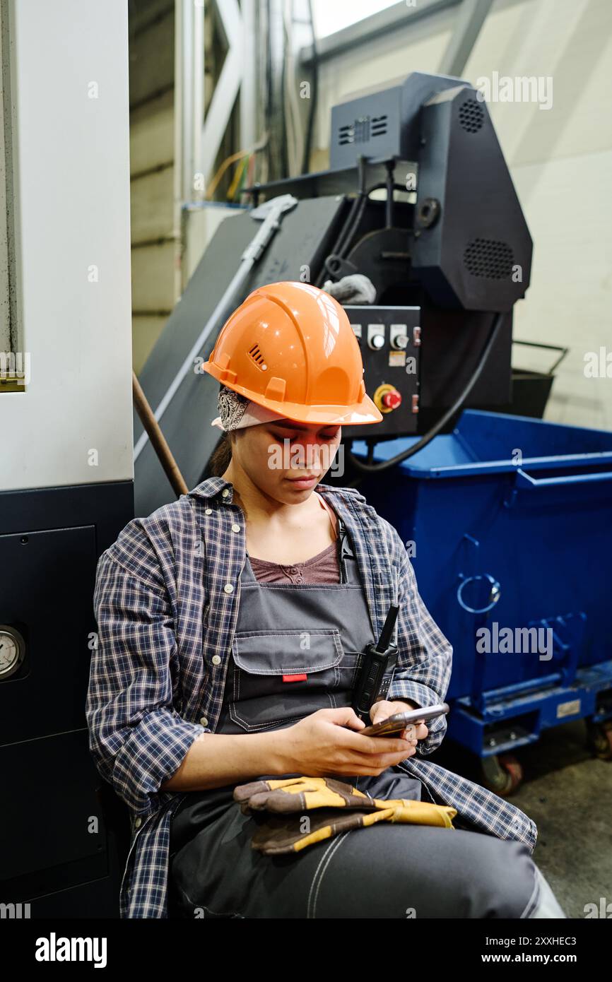 Donna che indossa un casco protettivo arancione e un'uniforme che si impegna con lo smartphone mentre si trova in uno stabilimento di produzione, mostrando concentrazione e concentrazione nell'ambiente industriale Foto Stock
