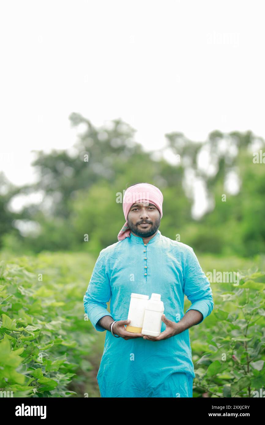 Felice contadino villaggio indiano che detiene la bottiglia di fertilizzante guardando la fotocamera a terreno agricolo - concetto di promozione del prodotto, raccomandazione e agricolo Foto Stock