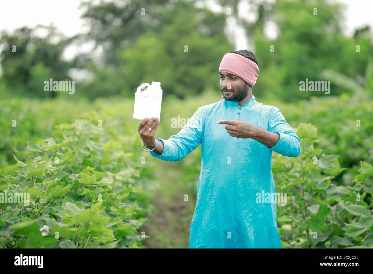 Felice contadino villaggio indiano che detiene la bottiglia di fertilizzante guardando la fotocamera a terreno agricolo - concetto di promozione del prodotto, raccomandazione e agricolo Foto Stock