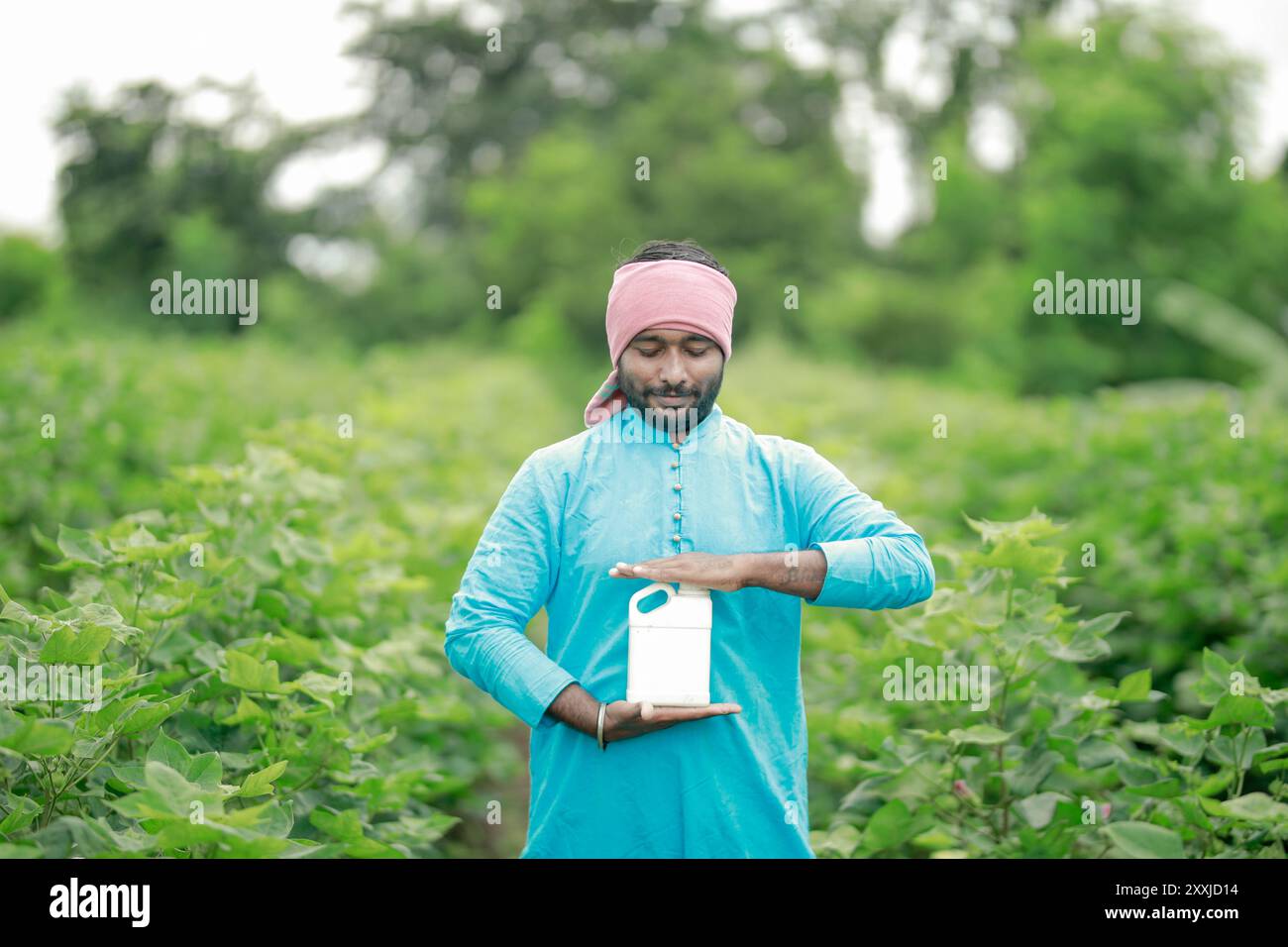 Felice contadino villaggio indiano che detiene la bottiglia di fertilizzante guardando la fotocamera a terreno agricolo - concetto di promozione del prodotto, raccomandazione e agricolo Foto Stock