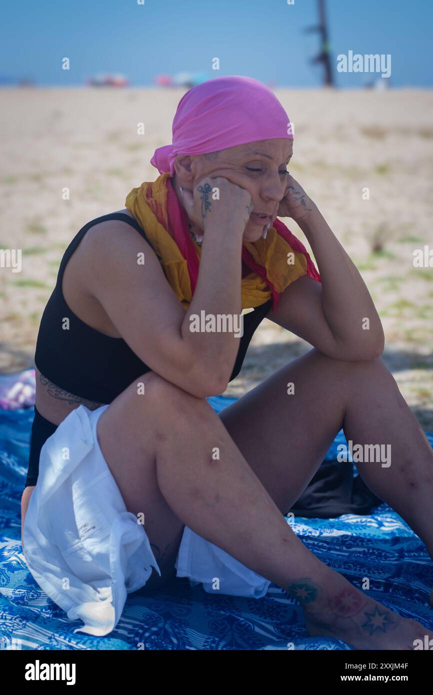 Sopravvissuto al cancro in una rimessa ponderata su una coperta da spiaggia, guardando in lontananza, incarnando la resilienza Foto Stock