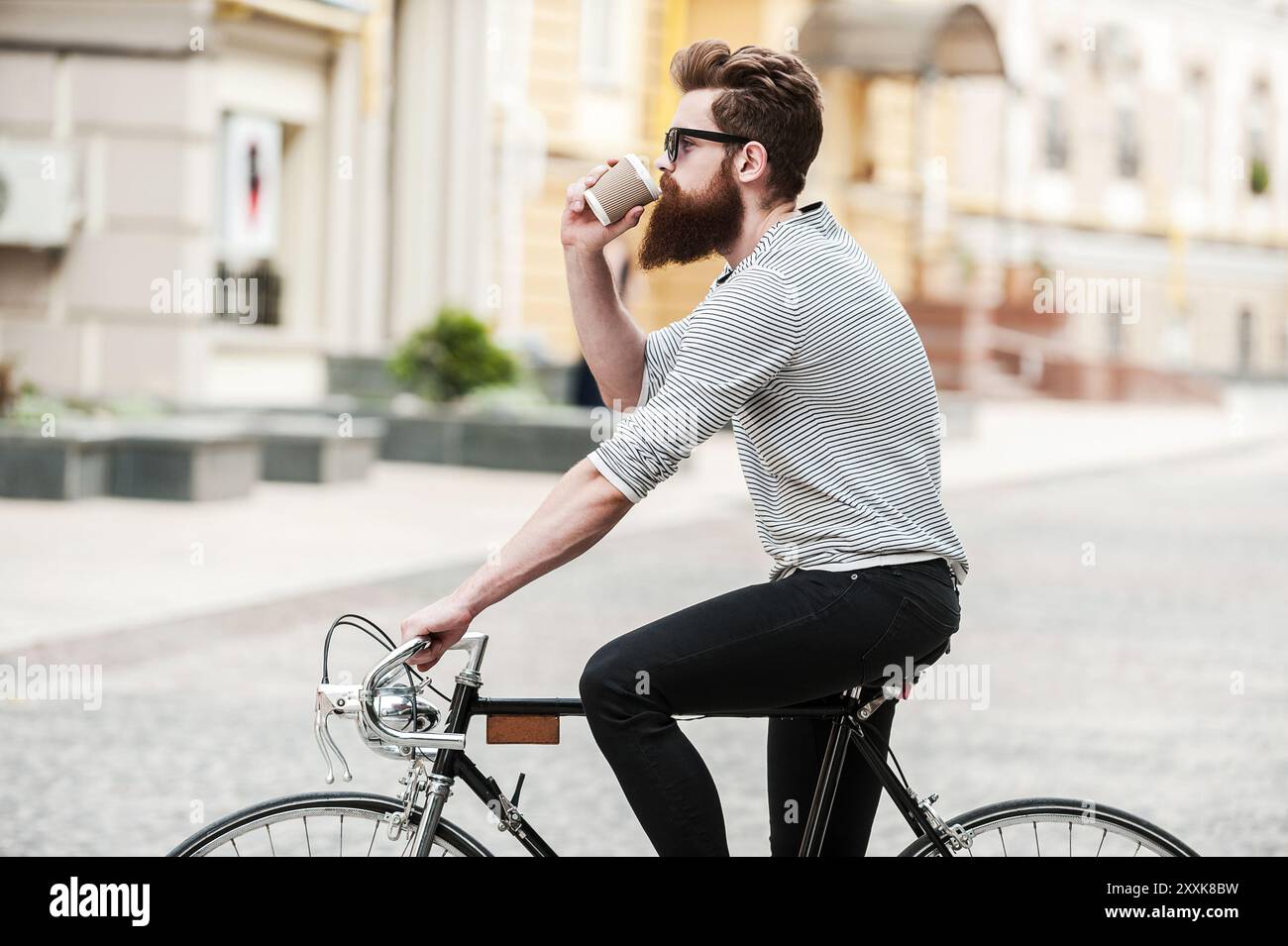 Caffè in movimento. Vista laterale di un giovane uomo bearded che beve caffè mentre si siede sulla sua bicicletta all'aperto Foto Stock