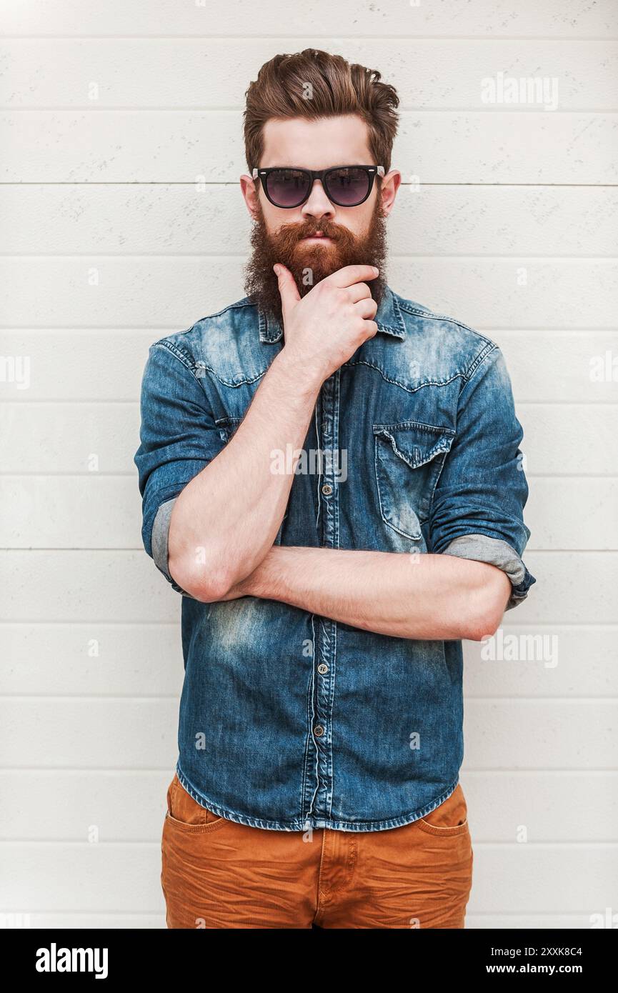 Fresco e sicuro di sé. Giovane uomo barbuto che guarda la macchina fotografica e tiene la mano sul mento mentre si trova all'aperto Foto Stock